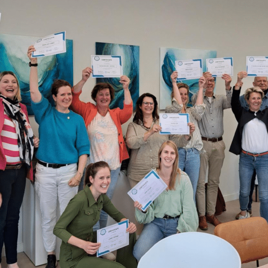 Groep mensen die glimlachen en certificaten vasthouden in een kamer.